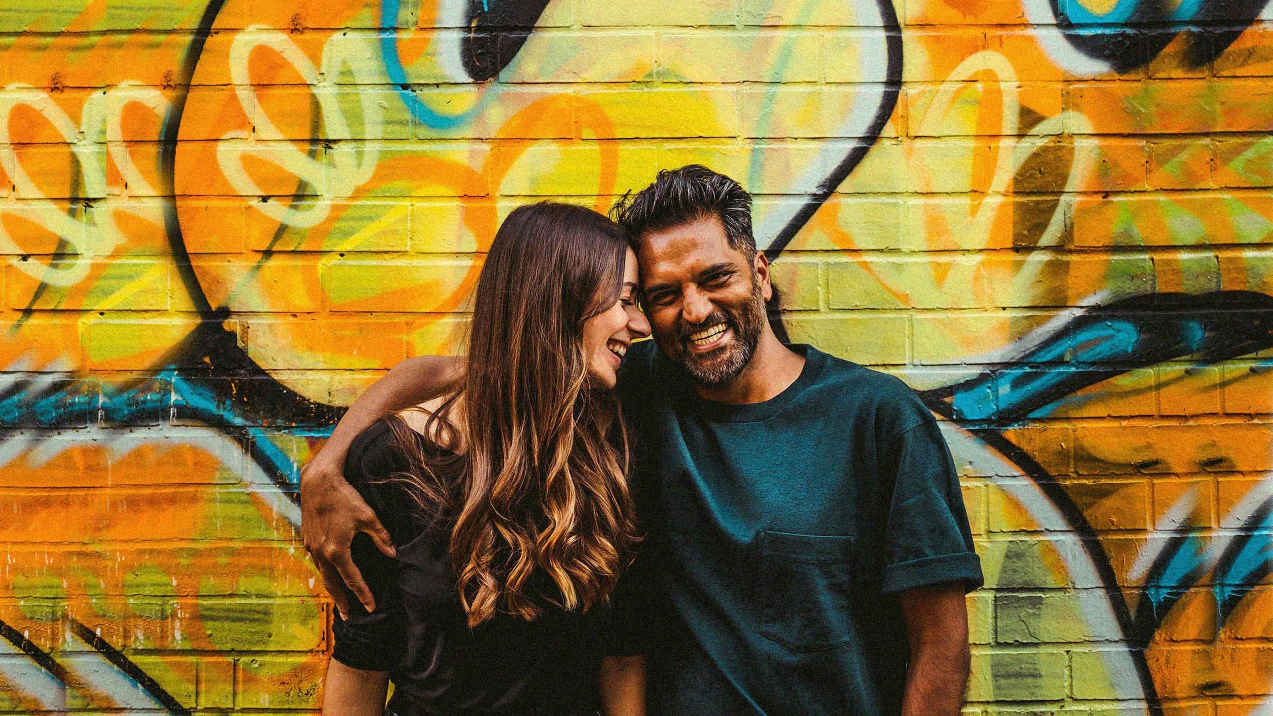 A man and lady stand in front of a multi coloured wall and pose for a photograph as the lady places her forehead onto the side of the man's head